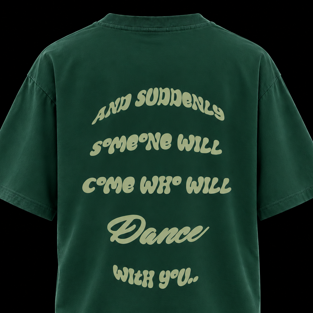 A dark green oversized t-shirt shown from the back, featuring a light green retro-style text graphic that reads 'AND SUDDENLY SOMEONE WILL COME WHO WILL Dance WITH YOU.' against a solid black background.