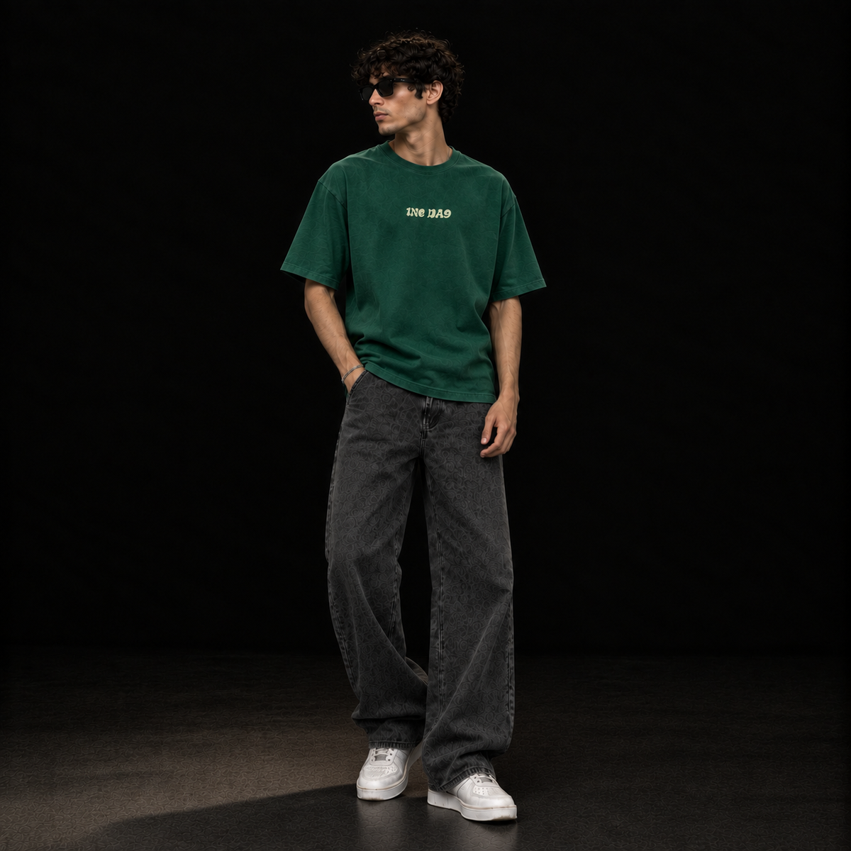 A man wearing a dark green oversized t-shirt with 'ONE DAY' printed in yellow on the chest, paired with dark gray wide-leg jeans and white sneakers, standing against a solid black background.