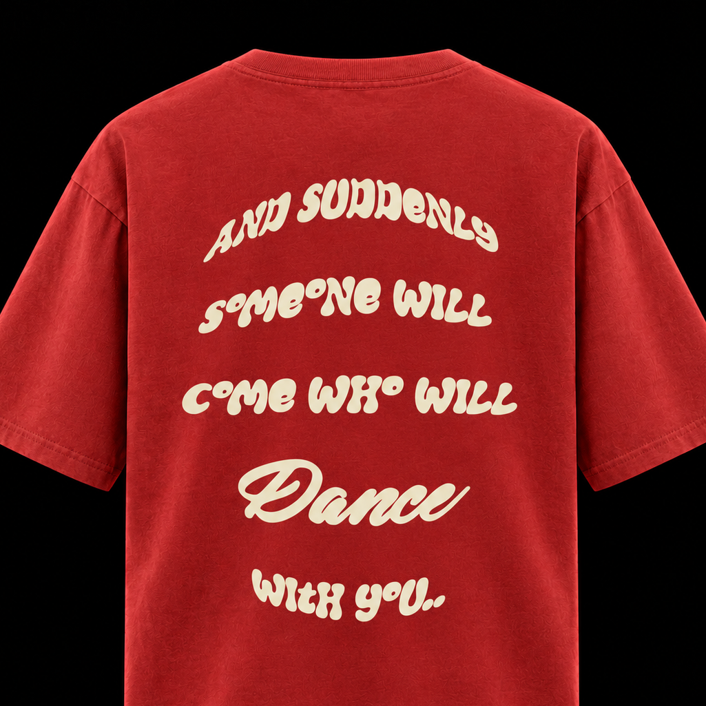 A red short-sleeve t-shirt shown from the back, featuring a large cream-colored retro-style text graphic that reads 'AND SUDDENLY SOMEONE WILL COME WHO WILL Dance WITH YOU'. The t-shirt is photographed against a solid black background.