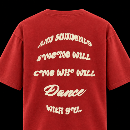 A red short-sleeve t-shirt shown from the back, featuring a large cream-colored retro-style text graphic that reads 'AND SUDDENLY SOMEONE WILL COME WHO WILL Dance WITH YOU'. The t-shirt is photographed against a solid black background.